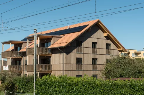 Aussenansicht eines mehrgeschossigen Wohnhauses in Holzbauweise mit Satteldach und integrierten Solarpanels. Die Fassade besteht aus senkrechter Holzschalung, die Balkone sind aus Holz gefertigt. Das Gebäude steht in einem Wohnquartier mit Hecken und Nachbarbauten, im Vordergrund verlaufen Bahnoberleitungen vor blauem Himmel.