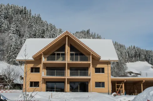 Aussenansicht eines neu gebauten Mehrfamilienhauses in Holzbauweise mit Satteldach und Balkonen im Winter. Das Gebäude steht in verschneiter Landschaft neben einem landwirtschaftlichen Anbau, umgeben von Hügeln und verschneiten Bäumen unter blauem Himmel.