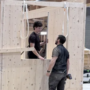 Zwei Mitarbeitende der Zimmerei Hirschi montieren in der Werkhalle ein vorgefertigtes Holzelement. Sie arbeiten an einer massiven Holzrahmenkonstruktion mit Fensteröffnung und stimmen sich dabei direkt vor Ort ab.