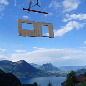 Vorgefertigtes Holzwandelement mit Fenster- und Türöffnung wird von einem Kran über einer alpinen Berglandschaft transportiert. Im Hintergrund sind grüne Berghänge und ein See zu sehen, welche die präzise Montage von Holzbauelementen in anspruchsvollem Gelände veranschaulichen.