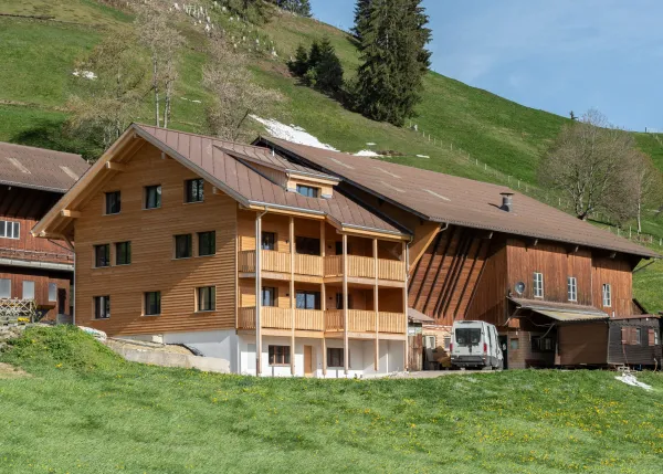 Aussenansicht eines neu errichteten Wohnhauses in Holzbauweise mit Satteldach und Balkonen, eingebettet in eine hügelige, ländliche Landschaft. Das Gebäude steht neben bestehenden landwirtschaftlichen Bauten. Im Vordergrund eine Wiese, im Hintergrund ein begrünter Hang mit Bäumen unter blauem Himmel.