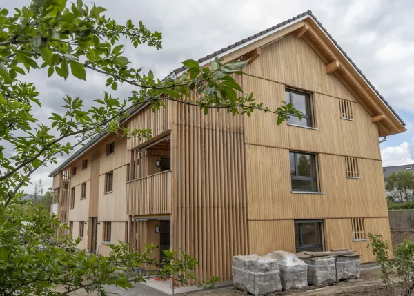 Aussenansicht eines neu errichteten Mehrfamilienhauses in moderner Holzbauweise mit senkrechter Holzfassade und Satteldach. Balkone und Fassadenelemente sind aus Holz gestaltet. Im Vordergrund sind Pflanzen und Baustellenmaterialien zu sehen, im Hintergrund ein Wohnquartier unter bewölktem Himmel.