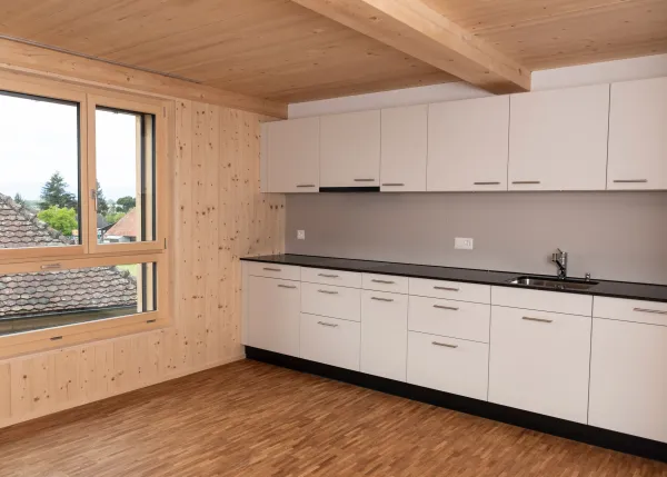 Innenansicht eines hellen Wohnraums in Holzbauweise mit sichtbarer Holzdecke und Holzboden. Grosse, bodentiefe Schiebetüren aus Glas führen auf einen überdachten Balkon mit Holzgeländer. Durch die Fenster fällt viel Tageslicht ein und ermöglicht den Blick nach draussen.