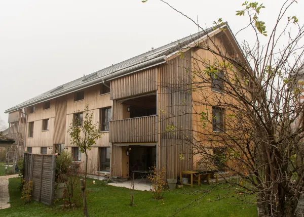 Aussenansicht eines neu errichteten Mehrfamilienhauses in moderner Holzbauweise mit senkrechter Holzfassade und Satteldach. Balkone und Fassadenelemente sind aus Holz gestaltet. Im Vordergrund sind Pflanzen und Baustellenmaterialien zu sehen, im Hintergrund ein Wohnquartier unter bewölktem Himmel.