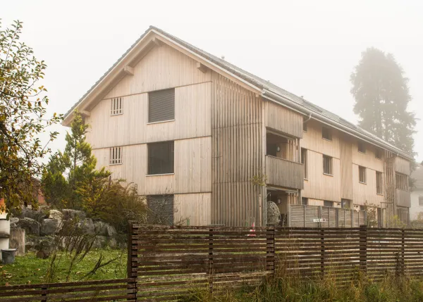 Aussenansicht eines neu errichteten Mehrfamilienhauses in moderner Holzbauweise mit senkrechter Holzfassade und Satteldach. Balkone und Fassadenelemente sind aus Holz gestaltet. Im Vordergrund sind Pflanzen und Baustellenmaterialien zu sehen, im Hintergrund ein Wohnquartier unter bewölktem Himmel.