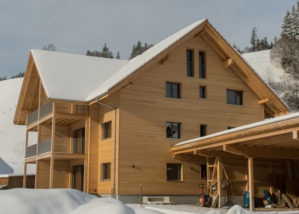 Aussenansicht eines neu gebauten Mehrfamilienhauses in Holzbauweise mit Satteldach und Balkonen im Winter. Das Gebäude steht in verschneiter Landschaft neben einem landwirtschaftlichen Anbau, umgeben von Hügeln und verschneiten Bäumen unter blauem Himmel.