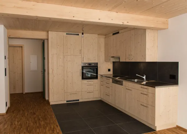 Heller Innenraum eines neu gebauten Holzhauses mit sichtbaren Holzbalken, offener Küche aus Holz, Treppenaufgang und grossen Fenstern mit Zugang zum Balkon. Der Raum wirkt modern, offen und natürlich durch die Kombination von Holz und klaren Linien.