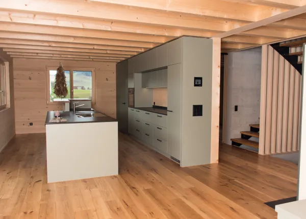 Innenansicht eines modernen Holzbaus mit sichtbaren Balken, natürlichen Holzoberflächen und massgefertigten Einbauten. Grosszügige Fensterflächen sorgen für viel Tageslicht und unterstreichen die warme, ruhige Wohnatmosphäre. Hochwertige Materialien und präzise Schreinerarbeiten prägen das Gesamtbild.