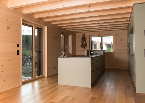 Innenansicht eines modernen Holzbaus mit sichtbaren Balken, natürlichen Holzoberflächen und massgefertigten Einbauten. Grosszügige Fensterflächen sorgen für viel Tageslicht und unterstreichen die warme, ruhige Wohnatmosphäre. Hochwertige Materialien und präzise Schreinerarbeiten prägen das Gesamtbild.