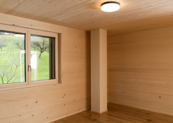 Innenansicht eines modernen Holzbaus mit sichtbaren Balken, natürlichen Holzoberflächen und massgefertigten Einbauten. Grosszügige Fensterflächen sorgen für viel Tageslicht und unterstreichen die warme, ruhige Wohnatmosphäre. Hochwertige Materialien und präzise Schreinerarbeiten prägen das Gesamtbild.