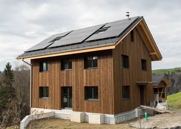 Neubau eines modernen Holzhauses am Hang mit dunkler Holzfassade, Balkon und grosser Fensterfront. Das Gebäude steht in ländlicher Umgebung mit Wiese und weiteren Häusern im Hintergrund unter bewölktem Himmel.