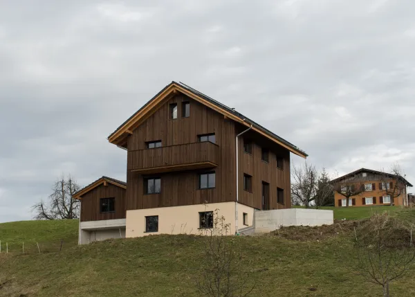 Neubau eines modernen Holzhauses am Hang mit dunkler Holzfassade, Balkon und grosser Fensterfront. Das Gebäude steht in ländlicher Umgebung mit Wiese und weiteren Häusern im Hintergrund unter bewölktem Himmel.