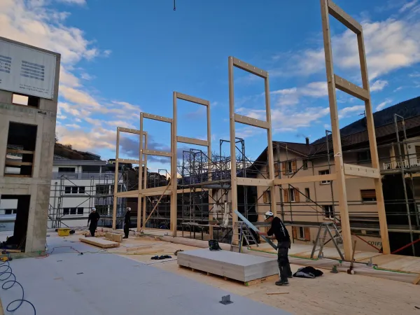 Zimmerleute arbeiten an der Holzrahmenkonstruktion eines mehrgeschossigen Gebäudes auf einer Baustelle. Die tragenden Wände aus Holz werden vor Ort montiert, während ein Baukran über dem Bauplatz steht. Im Hintergrund erstrecken sich Dorfhäuser und eine beeindruckende alpine Berglandschaft