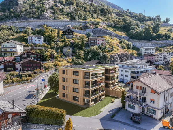 Visualisierung eines modernen Mehrfamilienhauses in Holzbauweise mit grossen Balkonen und begrüntem Dach mit Solaranlage. Das Gebäude fügt sich in eine alpine Dorflandschaft mit traditionellen Häusern, Strassen und einem bewaldeten Berghang im Hintergrund ein.