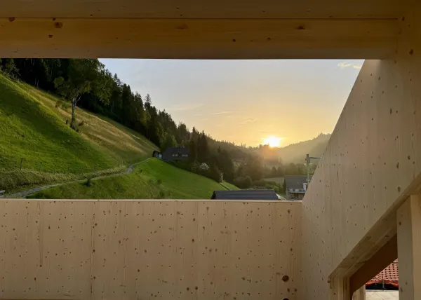Zimmerleute montieren während der Aufrichtearbeiten eine vorgefertigte Holzkonstruktion auf einem Gebäude. Ein Kran hebt ein grosses Dachelement an seinen Platz, während mehrere Fachkräfte auf dem Dach die Montage begleiten.